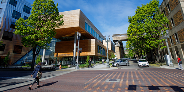 Modern urban street view with pedestrians crossing, cars parked, and a contemporary wooden building surrounded by green trees under a clear blue sky.