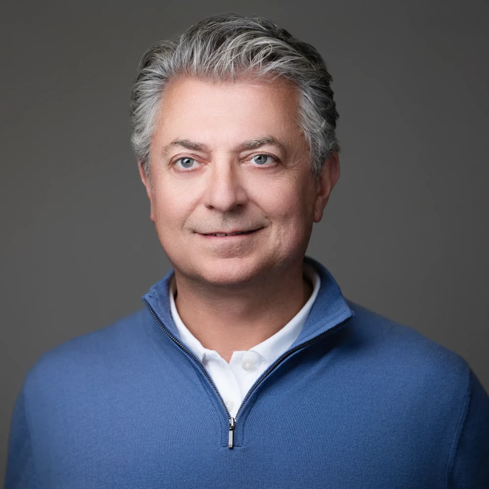 Professional headshot of CEO wearing a blue quarter-zip sweater over a white collared shirt against a neutral gray background.