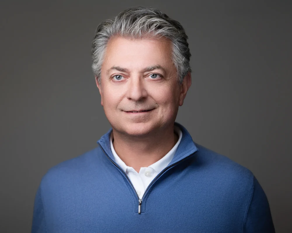Professional headshot of CEO wearing a blue quarter-zip sweater over a white collared shirt against a neutral gray background.