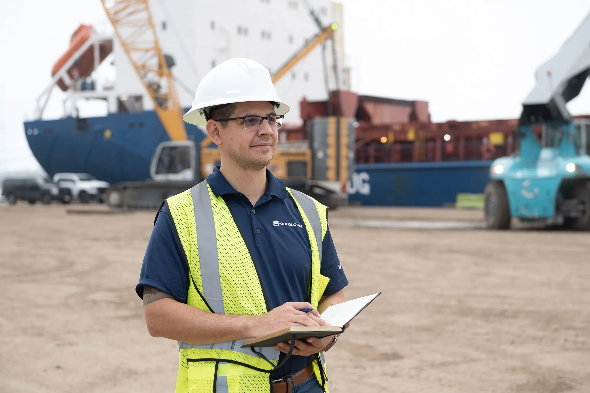 Port operations worker wearing a white hard hat and yellow safety vest holding a notebook in front of a cargo ship and heavy equipment.