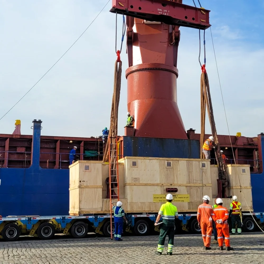 Heavy industrial cargo lifted by crane onto a flatbed trailer with workers supervising at a port.