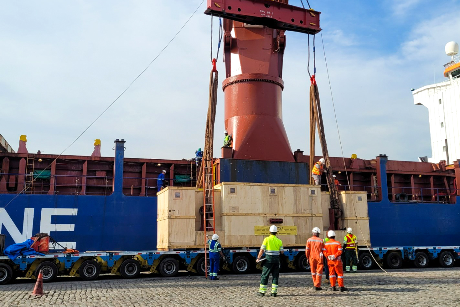 Trabalhadores do porto em pé ao redor de uma carga gigante que está sendo segurada por um guindaste
