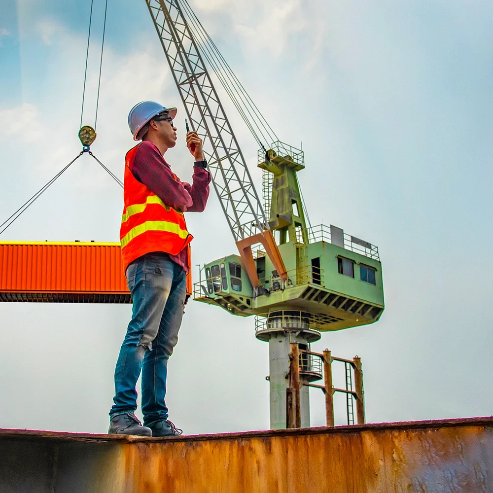 Worker using a handheld radio while supervising crane lifting a shipping container at a busy port terminal.