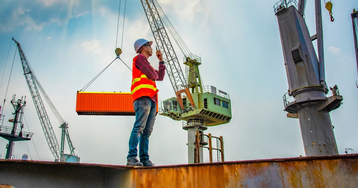 Worker using a handheld radio while supervising crane lifting a shipping container at a busy port terminal.
