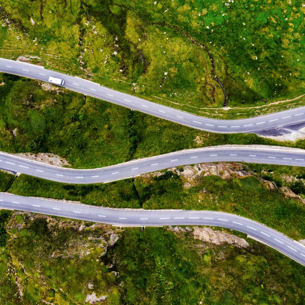 Aerial view of winding mountain road with sharp curves through green landscape and vehicles navigating the route.