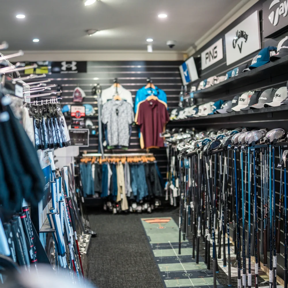 Interior of a golf retail store with racks of golf clubs, apparel, and branded caps displayed on shelves.