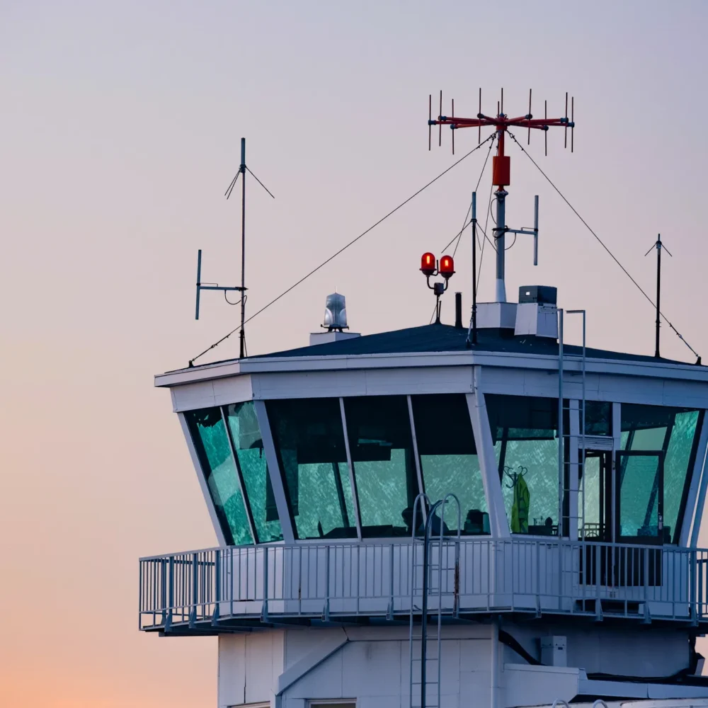 Airport control tower with antennas and communication equipment under a pastel-colored sunset sky.