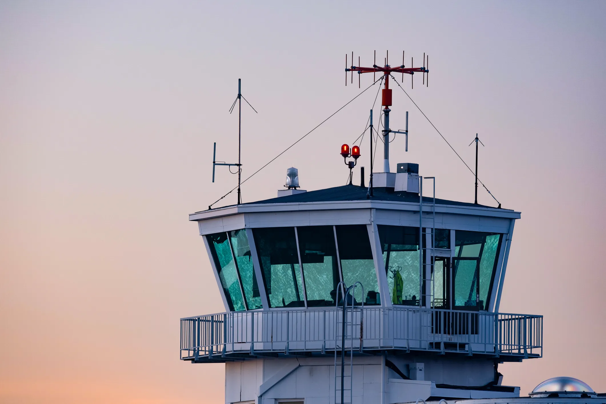 Torre de control del aeropuerto con antenas y equipos de comunicación bajo un cielo en tonos pastel al atardecer.
