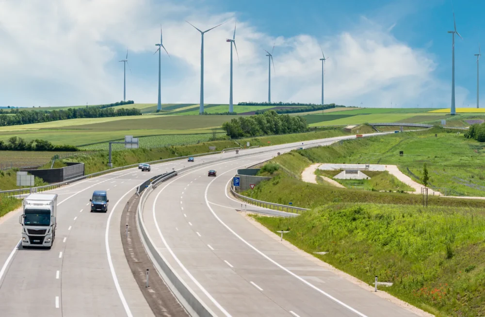 Highway with cars and a truck passing through green fields with multiple wind turbines in the background under a bright blue sky.