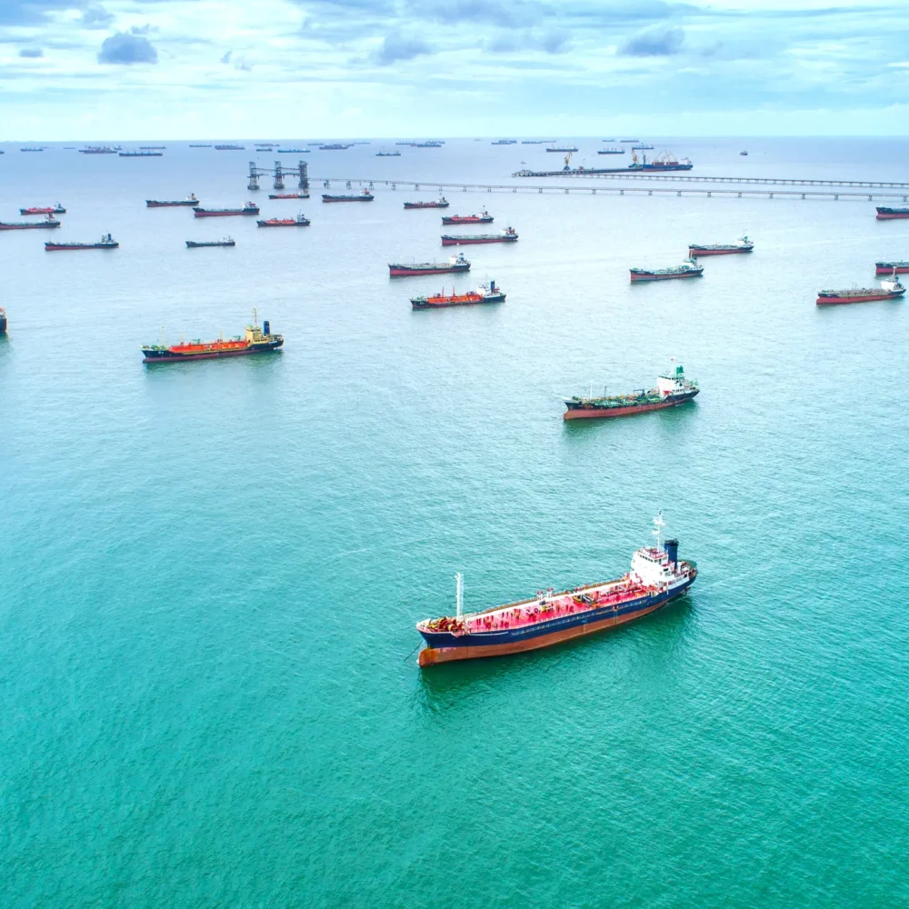 Aerial view of multiple cargo ships anchored in turquoise waters near an offshore oil terminal and long pier under a partly cloudy sky.