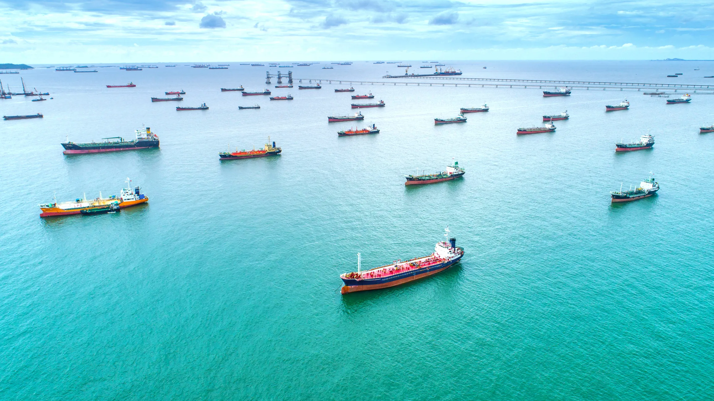Aerial view of multiple cargo ships anchored in turquoise waters near an offshore oil terminal and long pier under a partly cloudy sky.