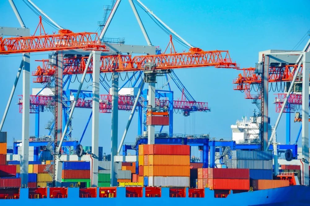 Colorful stacked shipping containers at a busy container terminal with multiple large orange gantry cranes under a clear blue sky.