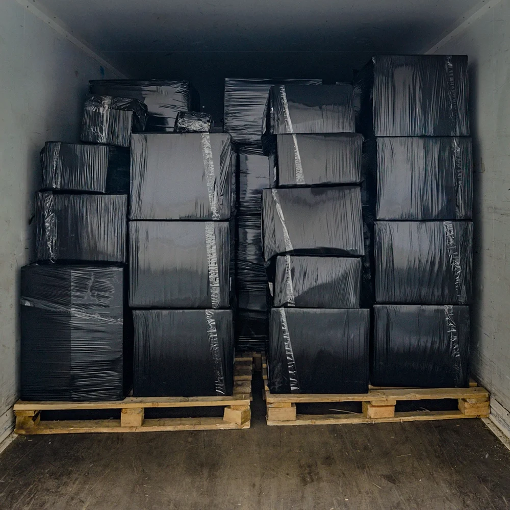 Stacked boxes wrapped in black plastic on pallets inside a shipping container for transport and distribution.