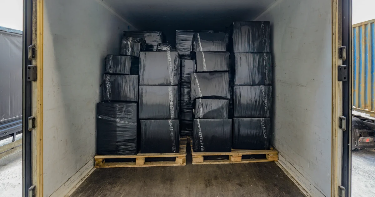 Stacked boxes wrapped in black plastic on pallets inside a shipping container for transport and distribution.