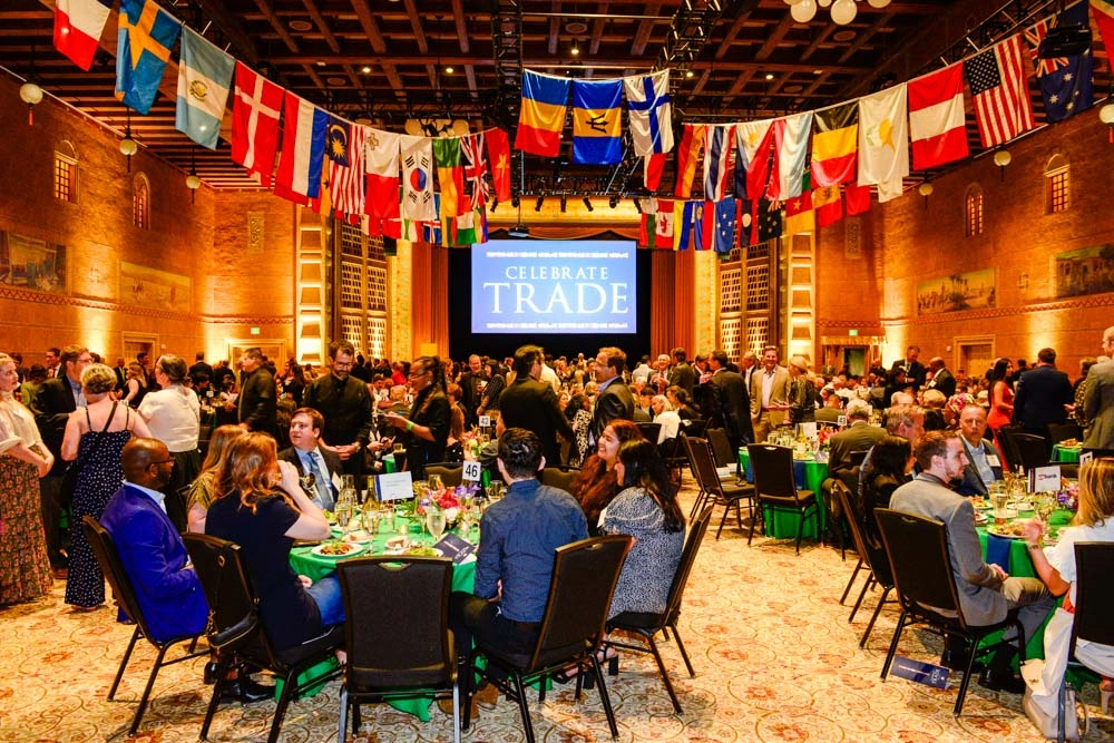 Large audience networking at Celebrate Trade event with tables, international flags, and stage backdrop.
