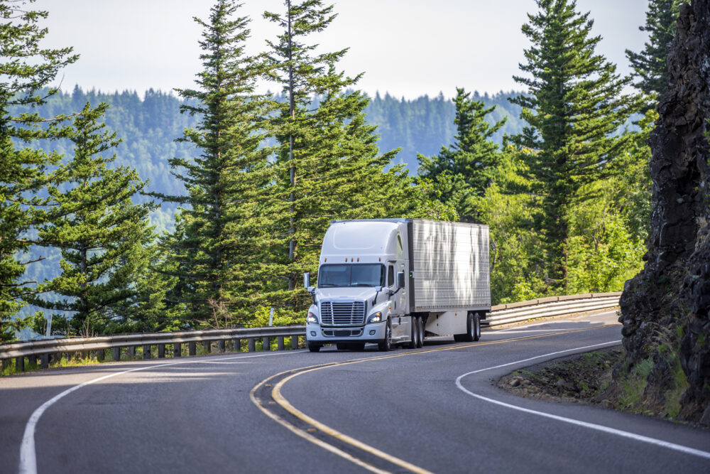 Big rig white powerful American bonnet long haul semi truck transporting commercial cargo in refrigerated semi trailer moving uphill on winding road with a green trees and safety fence on the side