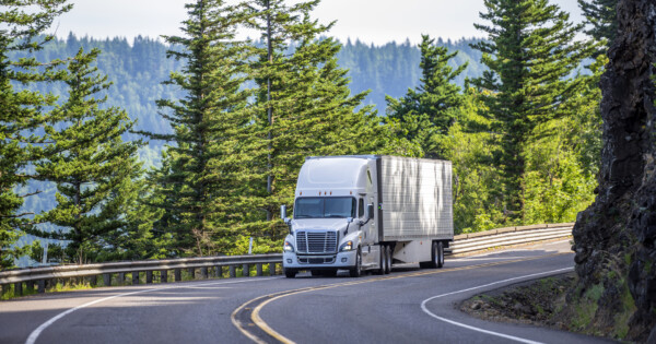 Big rig white powerful American bonnet long haul semi truck transporting commercial cargo in refrigerated semi trailer moving uphill on winding road with a green trees and safety fence on the side
