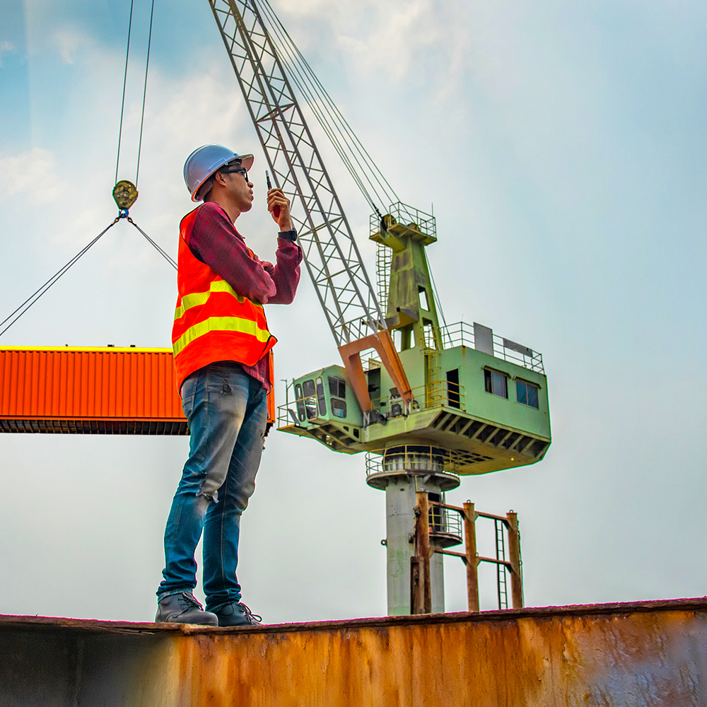 Trabajador portuario de pie sobre una estructura delante de una grúa en movimiento