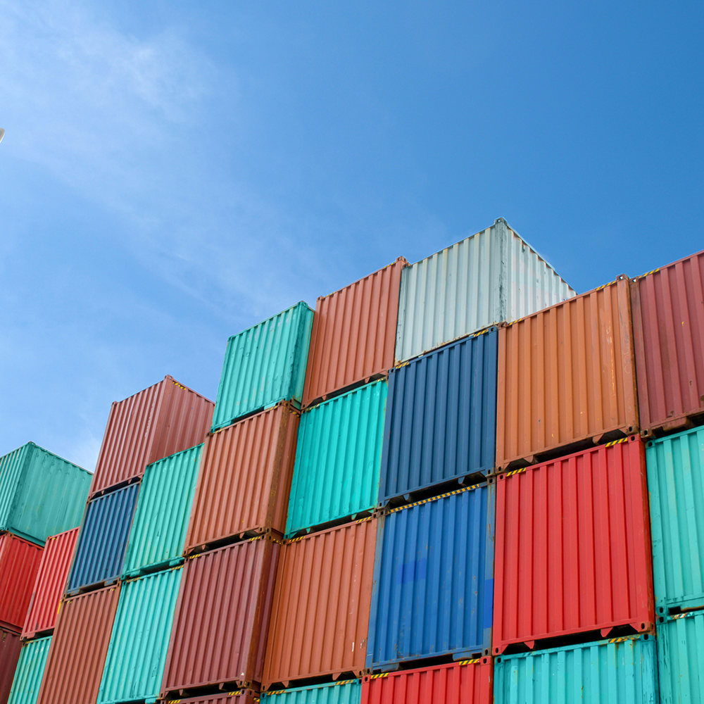 Plane flying over rows of stacked containers at a port.