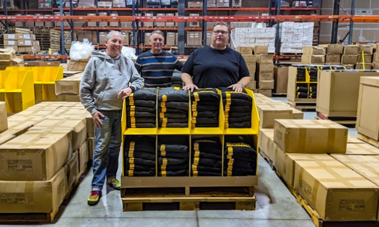 Team members standing behind a palletized display of DeWalt hoodies inside a warehouse filled with boxes and inventory.
