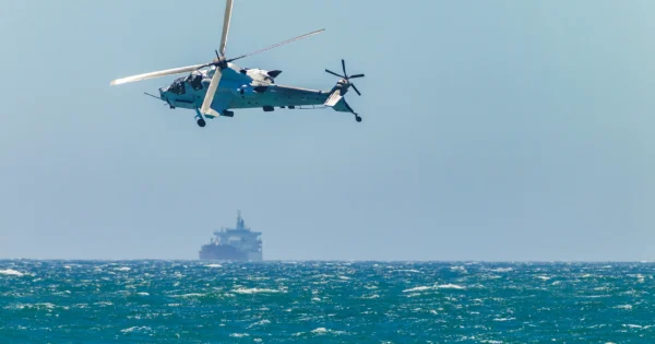 Military helicopter flying over the ocean with a cargo ship visible in the distant background under a clear blue sky.