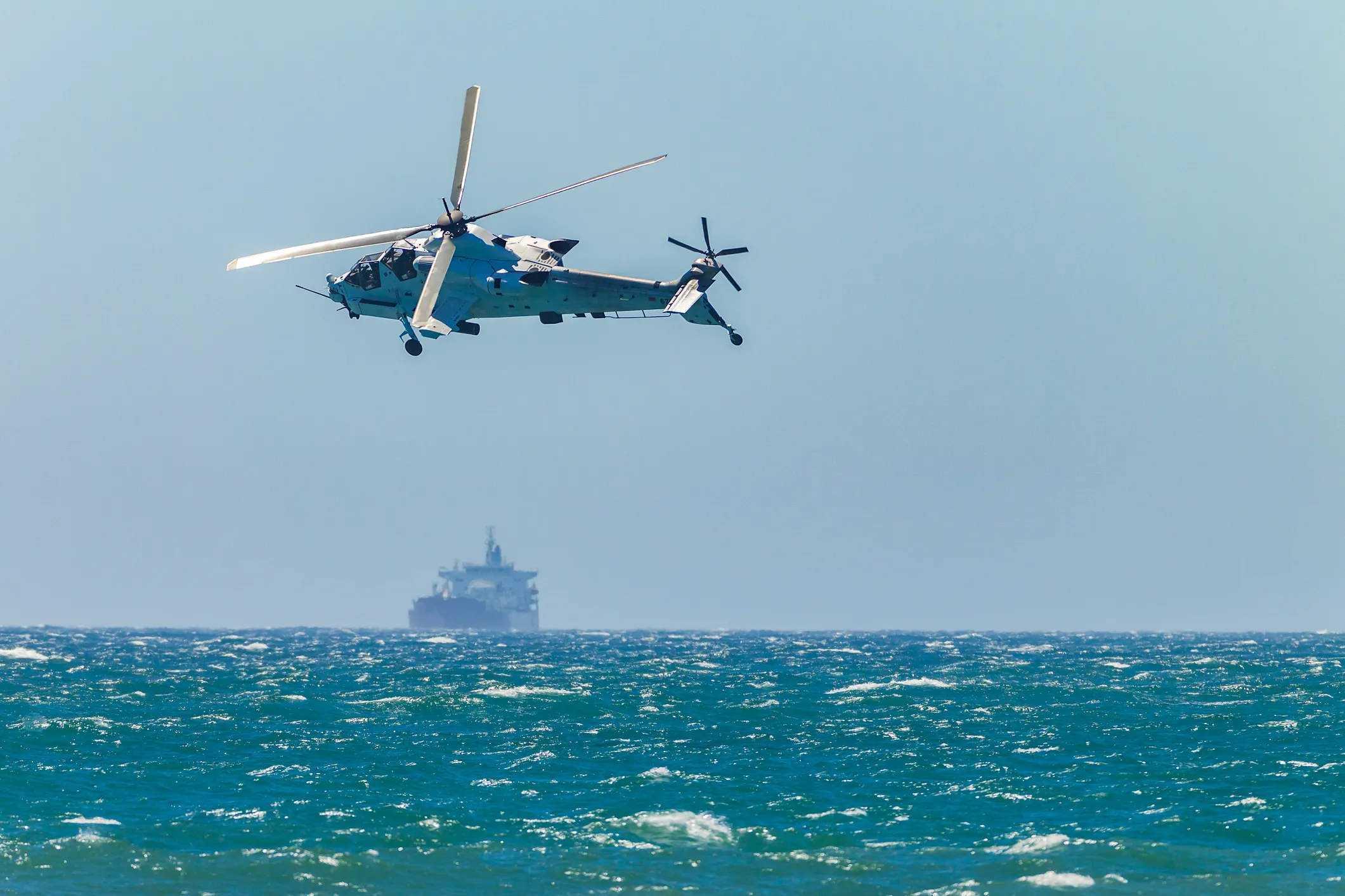 Military helicopter flying over the ocean with a cargo ship visible in the distant background under a clear blue sky.