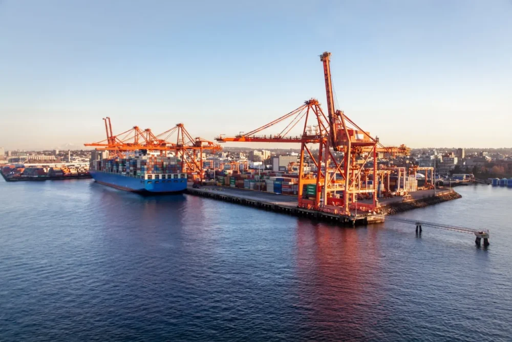Large cargo ship docked at a container terminal with towering orange cranes and stacked multicolored shipping containers under clear skies.