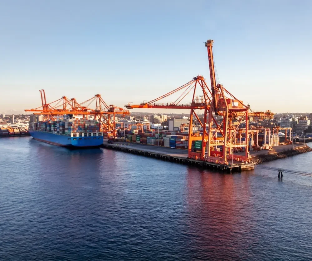 Large cargo ship docked at a container terminal with towering orange cranes and stacked multicolored shipping containers under clear skies.
