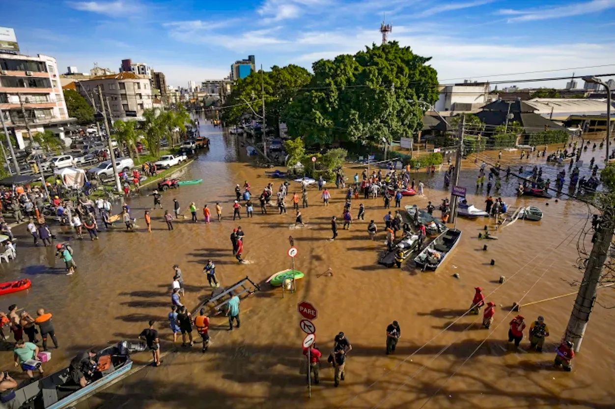 Flooded city street with people wading through water, rescue boats, and submerged vehicles after severe flooding in an urban area.