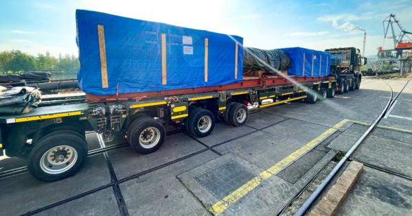 Oversized cargo wrapped in blue protective covering transported on a flatbed truck at an industrial port.