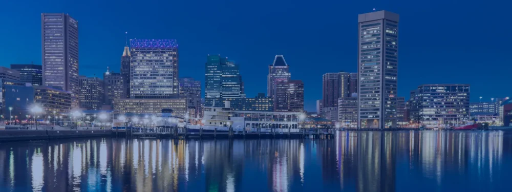 Baltimore city skyline at dusk with illuminated high-rise buildings reflecting on calm waterfront under a deep blue evening sky.