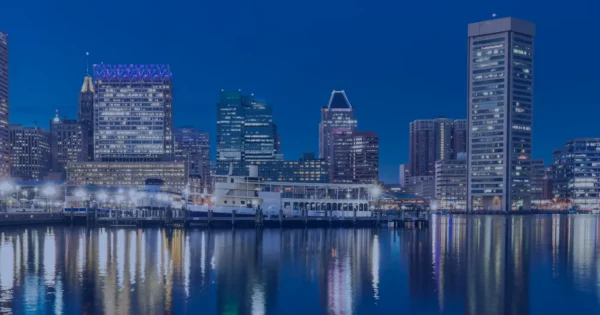 Baltimore city skyline at dusk with illuminated high-rise buildings reflecting on calm waterfront under a deep blue evening sky.