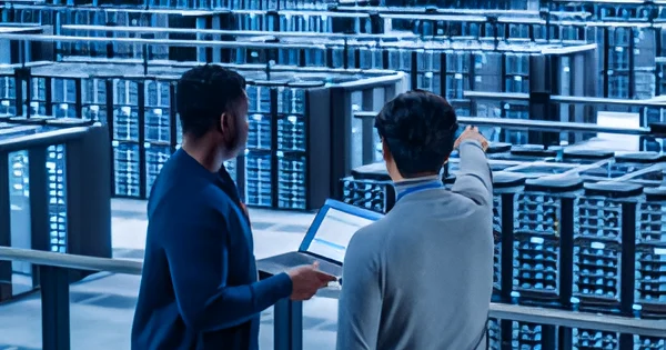 Two IT professionals standing in a large data center with rows of server racks, reviewing information on a laptop and pointing toward equipment.