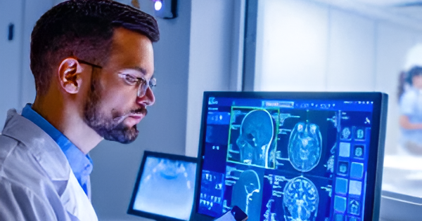 Healthcare professional reviewing brain scan images on a computer monitor while holding a tablet in a clinical setting.
