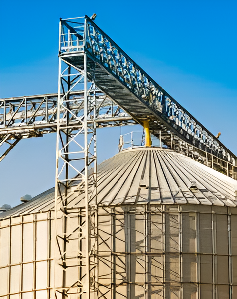 Industrial grain storage silo with metal roof and overhead conveyor system against a clear blue sky.