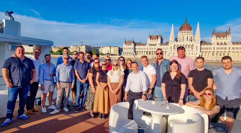OIA Global Hungary team group photo on a river cruise boat with the Hungarian Parliament building in Budapest in the background.