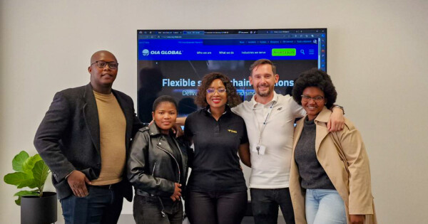 OIA Global team members standing together in office with company website displayed on screen behind them.