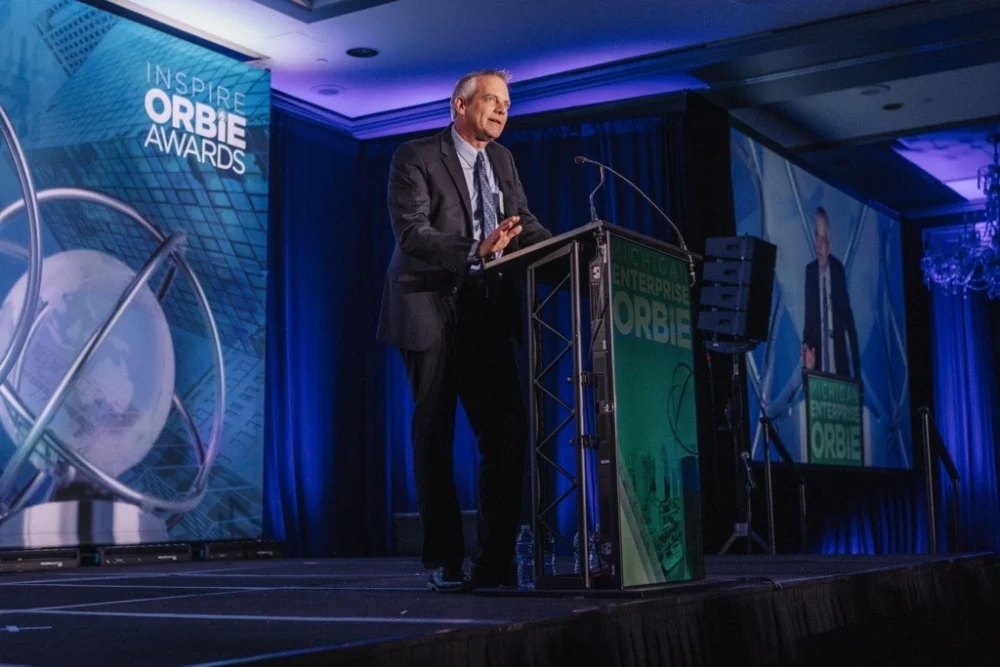 Business executive speaking at a podium during the Inspire ORBIE Awards event with branded stage backdrop.