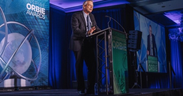 Business executive speaking at a podium during the Inspire ORBIE Awards event with branded stage backdrop.