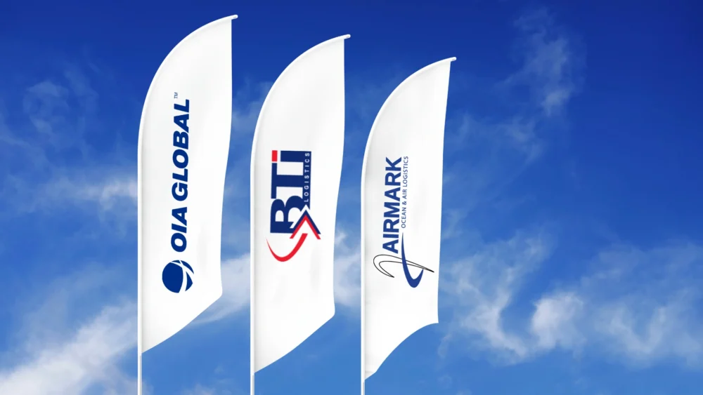 Flags displaying OIA Global, BTI Logistics, and Airmark logos against a blue sky representing logistics brand network.