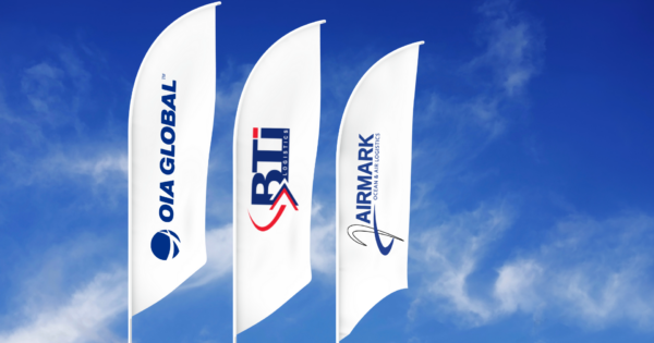 Flags displaying OIA Global, BTI Logistics, and Airmark logos against a blue sky representing logistics brand network.