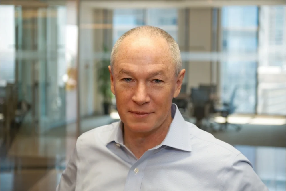 Professional corporate headshot of a business leader in an office environment with glass meeting rooms in the background.