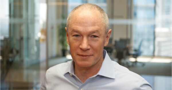 Professional corporate headshot of a business leader in an office environment with glass meeting rooms in the background.
