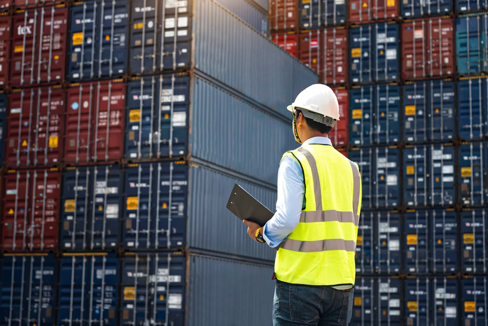 Person wearing a safety helmet and high-visibility vest inspecting stacked shipping containers at a port while holding a clipboard.