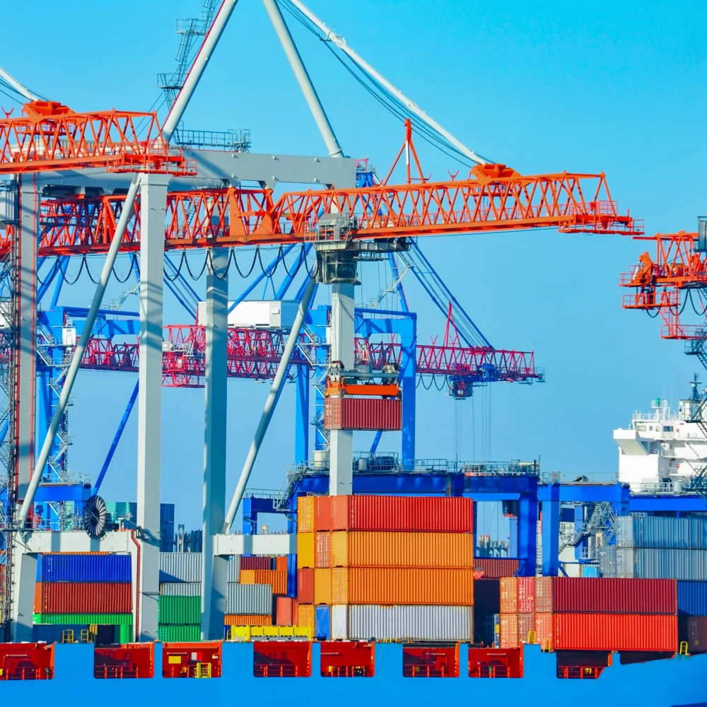 Colorful stacked shipping containers at a busy port with multiple large cargo cranes under a clear blue sky.