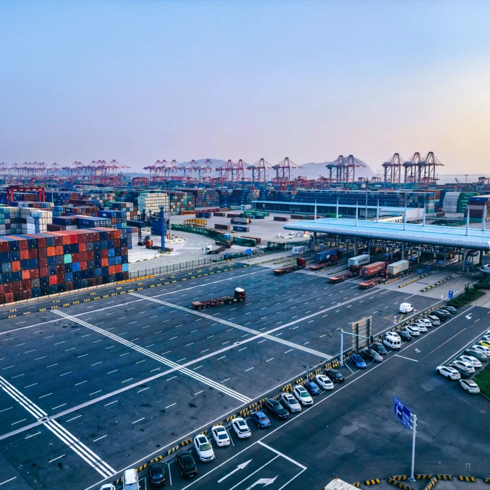 Aerial view of a large container port with stacked shipping containers, multiple cranes, trucks, and parking areas under a clear sky.