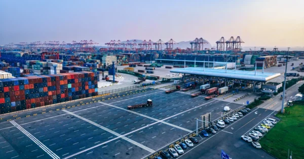 Vista aérea de um grande porto de contêineres com contêineres empilhados, vários guindastes, caminhões e áreas de estacionamento sob um céu claro.