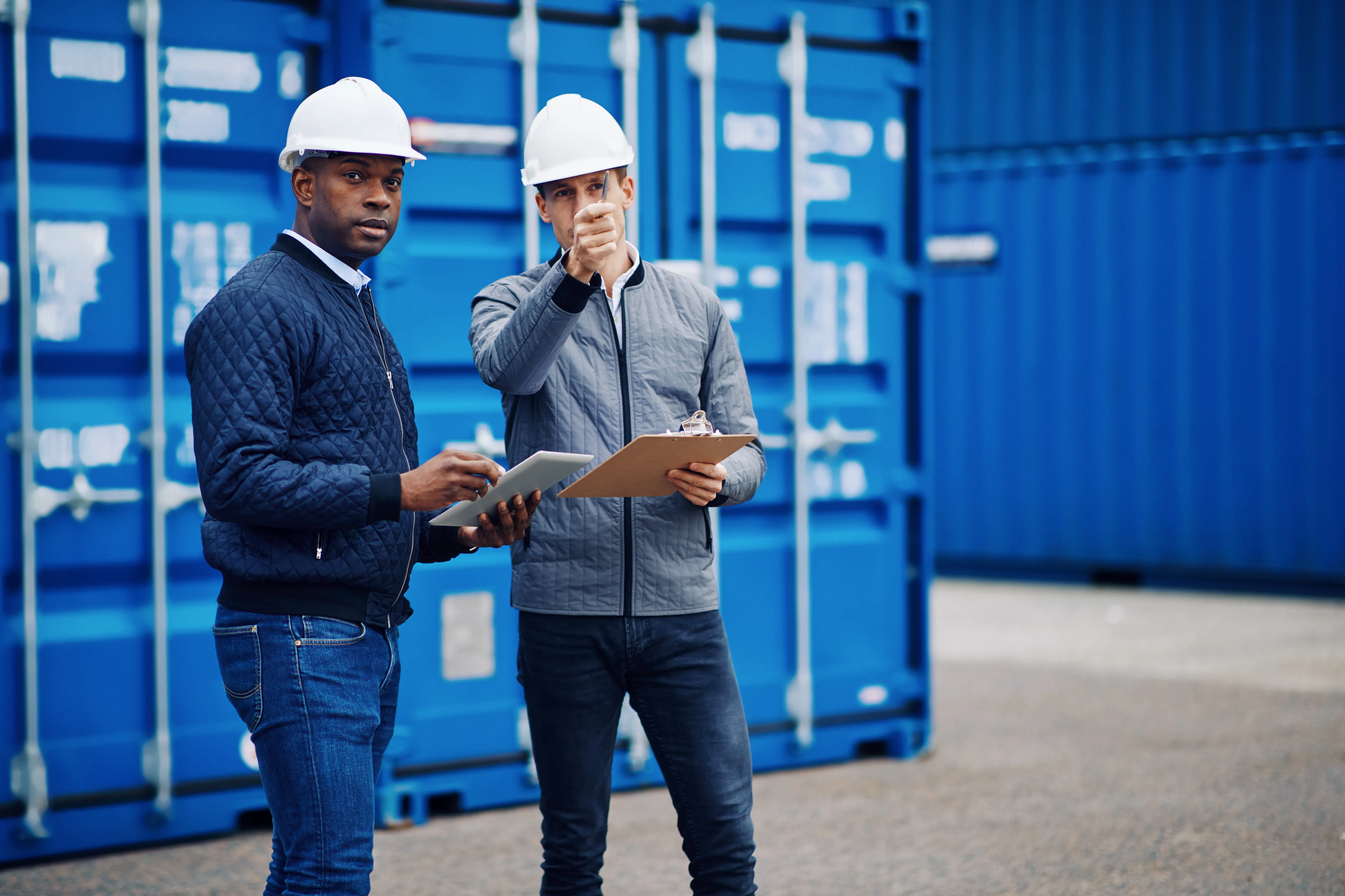 Two workers wearing safety helmets standing in front of blue shipping containers, reviewing documents on a clipboard and tablet at a logistics site.