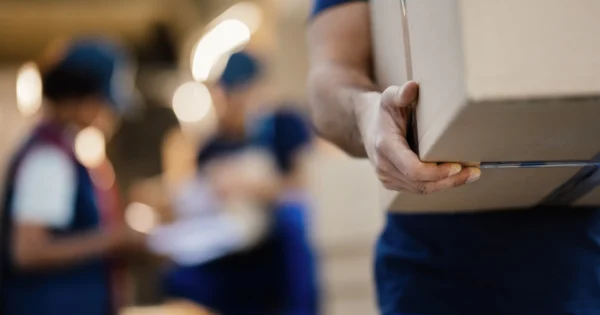 Close-up de uma pessoa carregando uma caixa de papelão em um armazém ou ambiente de distribuição, com outros trabalhadores e pacotes desfocados ao fundo.