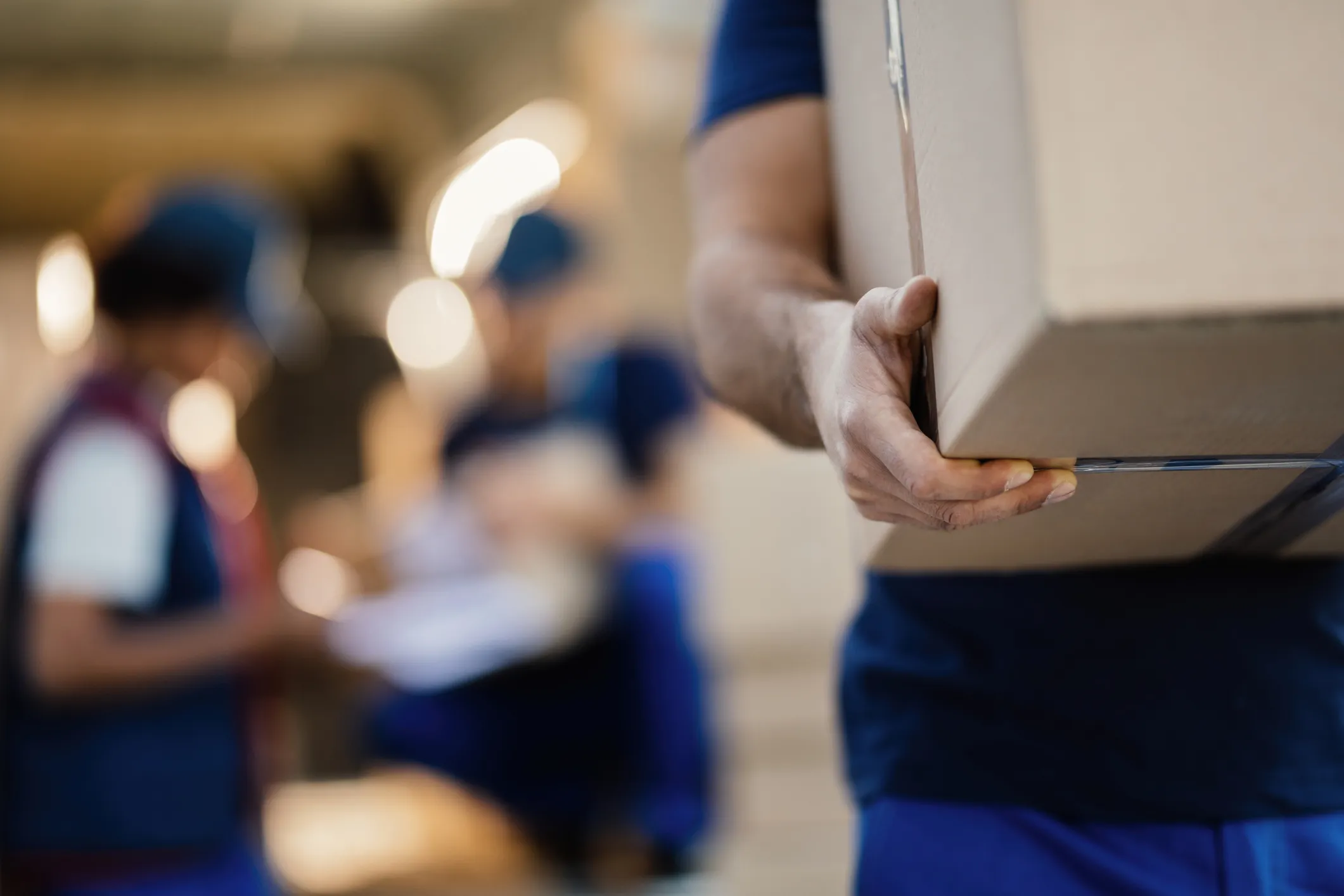 Close-up of a person carrying a cardboard box in a warehouse or distribution setting, with other workers and packages blurred in the background.